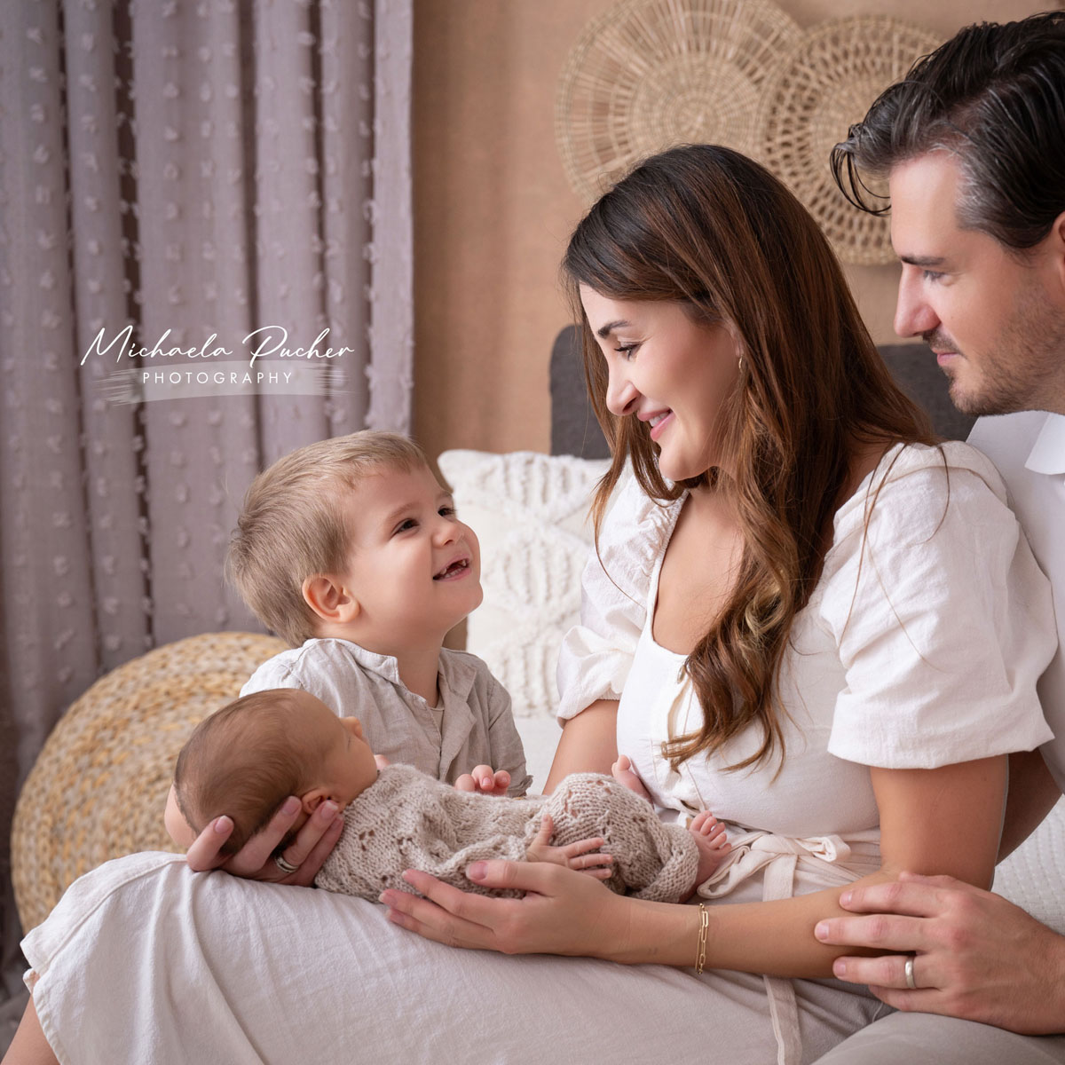 Familie sitzt eng beisammen, während das größere Kind lächelnd das Neugeborene betrachtet, das in den Armen der Mutter liegt. Der Vater sitzt liebevoll daneben.